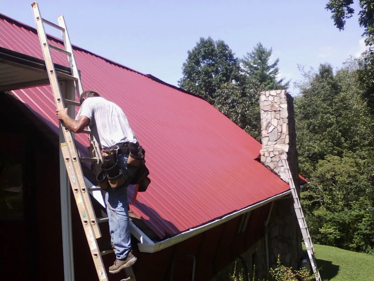 Roofer on a ladder beside a red metal roof and stone chimney — Soffit & Fascia Repair by Roofing Brothers in West End